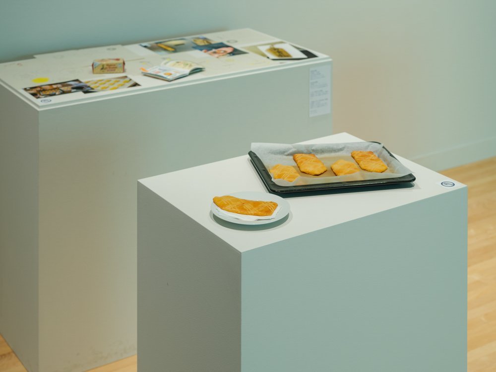 手前の小さな展示台には、細長い家のような形のクッキーが、１枚は白い皿に、４枚がオーブンの天板に置かれて展示されている。奥の大きな展示台には、写真やドローイング、本などが乱雑に置かれている。